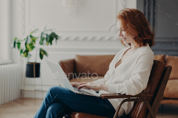 Woman freelancer sits in armchair, keyboards on laptop stock photo NULLED